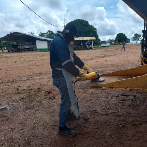 Operador afilando la pala de una retroexcavadora con esmeriladora angular durante mantenimiento preventivo de maquinaria pesada, usando equipo de protección personal.